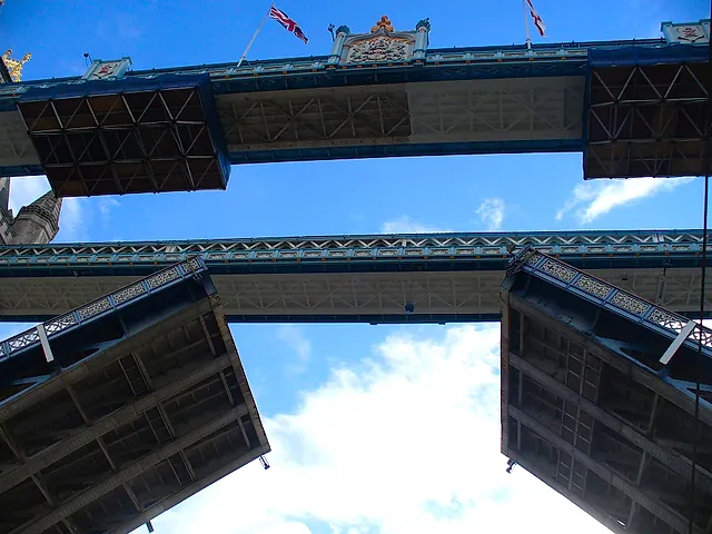 Tower Bridge with its two bascules raised, seen from below against a blue sky with a flag flying above